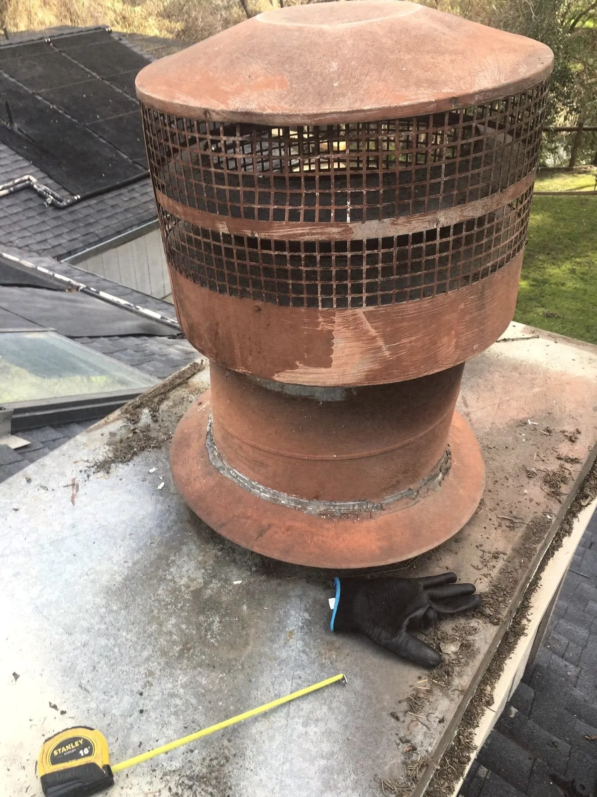 A clay pot chimney cap with a wire screen on a roof, next to a tape measure and gloves